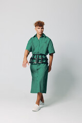 Fototapeta premium Young man walking in green shirt dress with black belt and white sneakers on plain background. Fashion model posing in casual outfit with curly hair and confident expression.