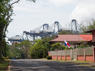 Obraz premium Massive gantry cranes tower over tropical port neighborhood with Panama flag and houses.