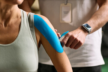Naklejka premium Caucasian young adult woman receiving kinesiology tape application on shoulder from Caucasian young adult man during physical therapy rehabilitation session in clinical setting