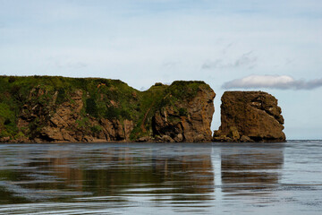 A dramatic rock formation with steep green cliffs splits at the shoreline, reflecting in the calm ocean waters beneath a bright sky. Tikhaya Bay in the Sea of Okhotsk, Sakhalin Island