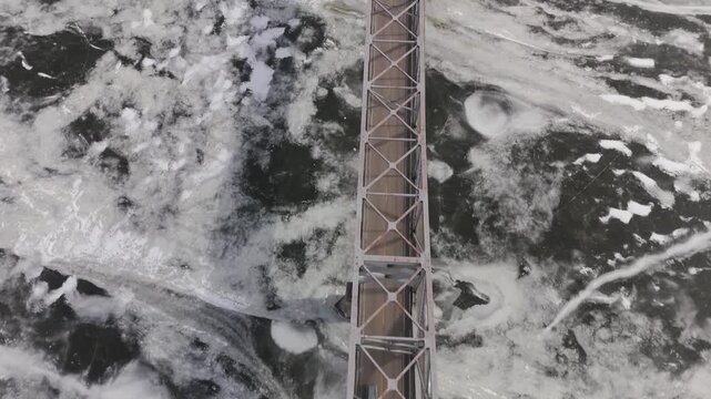 High-resolution aerial footage of the Mississippi River frozen during winter with a bridge crossing in Dubuque, Iowa. Vehicles pass overhead as ice formations create dramatic seasonal visuals.