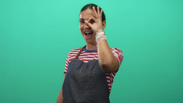 Woman making ok sign over her eye wearing denim apron and striped shirt in studio with teal backdrop; playful curiosity.