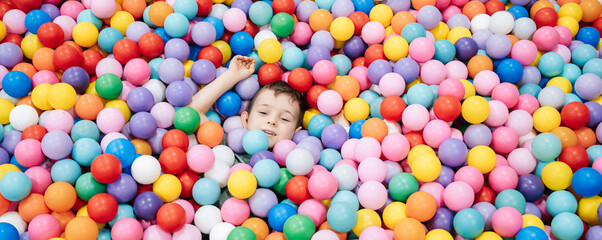 A happy little boy has fun jumping into a dry pool with colorful balls. The kid boy is playing and...