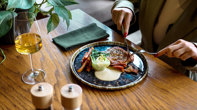 Close-up of a womans hands cutting a gourmet dish with knife and fork at a restaurant table, capturing fine dining rituals and relaxed lifestyle moments.