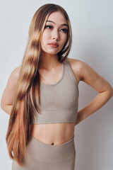 Fototapeta premium Young woman with long brown hair wearing beige sportswear posing with arms behind back against white background for fitness and wellness concept.