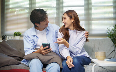 Obraz premium Happy couple sitting on a sofa and looking at a smartphone together, technology, and everyday life concept.