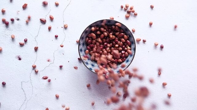 Slow motion pouring of raw peanuts into patterned ceramic bowl on white stone background with dynamic movement