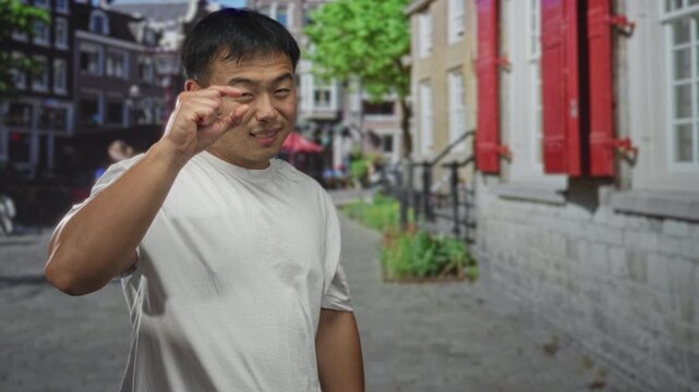 Man wearing white tshirt pinches thumb and forefinger on urban brick street beside shuttered building; playful precision.