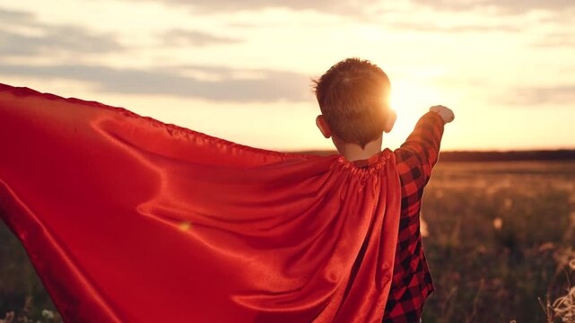 Playful boy stands in superhero character pose with red cape in field at sunset in evening. Boy with red cape imitates superhero flight. Fantasy of boy with red cape and superhero realization