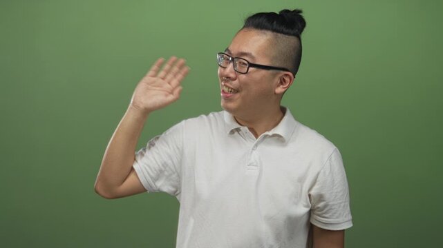 Man waving right hand and smiling while wearing glasses and a white polo shirt in a studio with a green backdrop; friendly greeting.