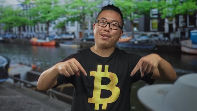 Man in bitcoin t shirt points both fingers down on street in amsterdam canal area; crypto enthusiasm confidence.