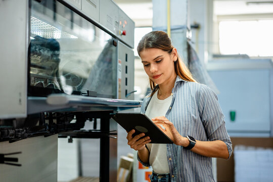 Female worker inspecting machinery in printing factory, using digital tablet for monitoring production and quality control. Concept of manufacturing process, technology, industrial, and efficiency.