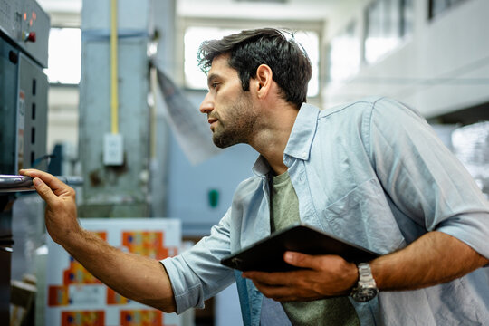 Professional male printer operator using a digital tablet to monitor quality control on an industrial printing press machine in a modern factory