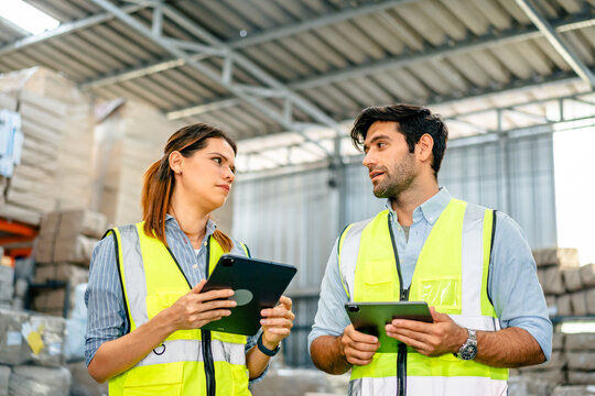 Diverse team of warehouse supervisors in safety vests using digital tablets to manage inventory and logistics in a large industrial printing facility storage area