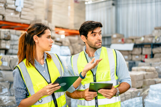 Diverse team of warehouse supervisors in safety vests using digital tablets to manage inventory and logistics in a large industrial printing facility storage area