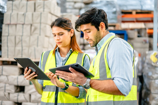 Diverse team of warehouse supervisors in safety vests using digital tablets to manage inventory and logistics in a large industrial printing facility storage area
