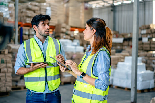 Diverse team of warehouse supervisors in safety vests using digital tablets to manage inventory and logistics in a large industrial printing facility storage area