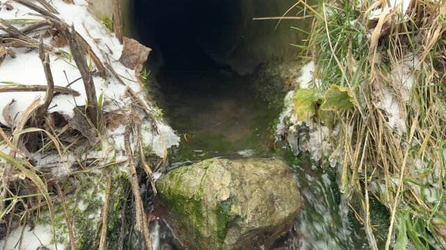 Water flows from a dark culvert and rushes over a moss-covered stone. The force of the stream against the rock produces a vibrant bubbling and a constant, refreshing splashing sound.