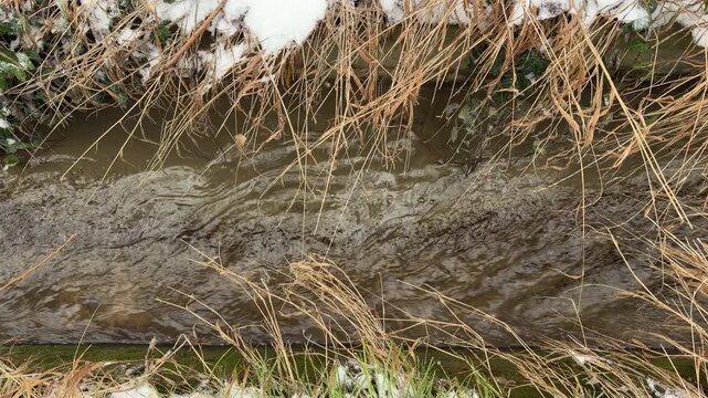 A steady stream of water rushes through a circular concrete culvert and out into a snowy channel. The movement of the water against the pipe and bank creates a resonant gurgling and splashing sound.
