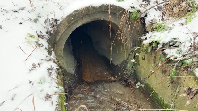 Water flows steadily through a dark concrete culvert into a snowy ditch. The liquid moves over the stone pipe, creating a hollow echoing trickle and a soft splashing sound.