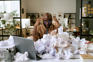 Black man sitting at cluttered desk surrounded by crumpled papers, holding head in hands, appearing...