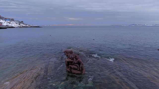 A close-up drone shot of the iconic Sedna (Sassuma Arnaa) bronze sculpture submerged in the tide of the Labrador Sea, set against a snowy coastline and twilight sky.