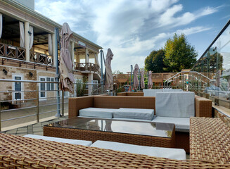 Obraz premium Empty outdoor seating area of resort restaurant terrace with stylish wicker furniture, comfortable cushions, glass-top coffee tables, and closed beige umbrellas under cloudy sky