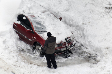 Adult male clearing deep accumulation of white snow from top and sides of red passenger car using shovel after heavy winter snowfall © HENADZY