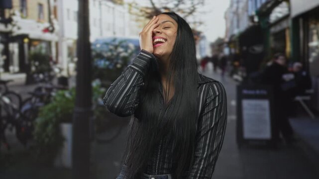Latin woman with long dark hair covering her face with her hand on street while laughing in a candid moment; happiness.