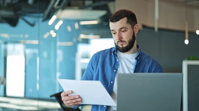Focused professional reviews documents and laptop in an office. Diligent and thoughtful, showing dedication to work. Reflects project management, data analysis, and concentrated effort.