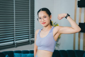 Young woman flexing arm muscle indoors showing strength and confidence with glasses and fitness watch in casual sportswear