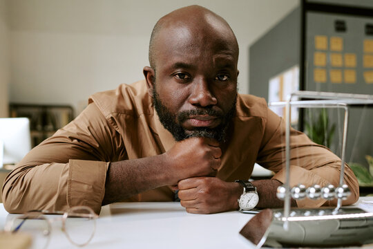 Portrait of Black man sitting at desk resting chin on hand looking into camera with serious expression, office environment visible in background business context