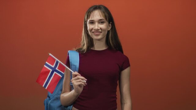 Young student woman holding norwegian flag and backpack strap, smiling with closed eyes in orange studio; pride celebration.