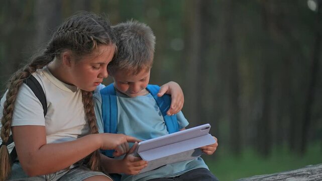 Outdoor tablet reading in forest. Boy and girl sit close as reader explores screen. Friend leans in, smile shared. Hiker backpack on child. Tablet focus for student and friend in forest afternoon.