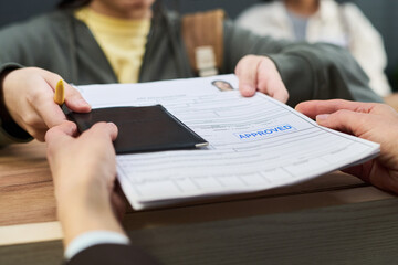 Plakat Girl handing passport and approved visa application form to unknown woman across counter at visa center, background showing blurred people waiting