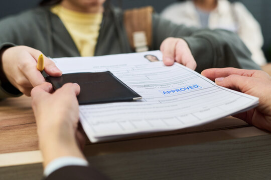 Girl handing passport and approved visa application form to unknown woman across counter at visa center, background showing blurred people waiting