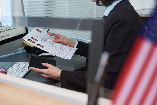 Middle aged Caucasian woman processing visa applications at visa center, holding passport and documents with applicant photos, sitting at desk with computer, American flag visible in foreground