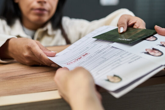 Woman handing over rejected visa application form and passport to another across counter at visa center, closeup of hands and documents visible