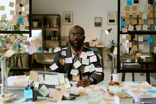 Portrait of Black man sitting at cluttered office desk covered with sticky notes, wearing glasses and suit, looking into camera, working on business tasks surrounded by reminders