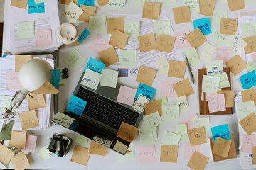 Overhead view of office desk showing laptop surrounded by scattered sticky notes, documents, lamp...