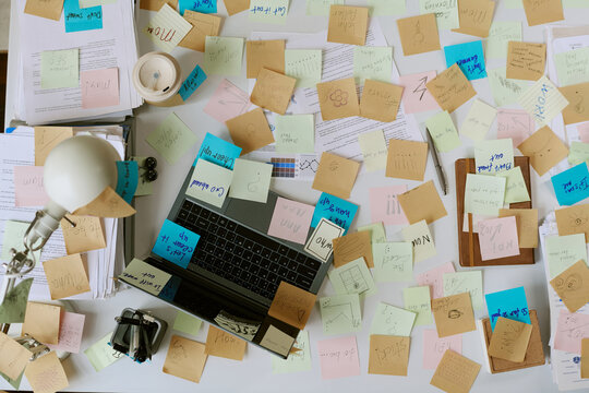Overhead view of office desk showing laptop surrounded by scattered sticky notes, documents, lamp and coffee cup, illustrating busy business planning and brainstorming process