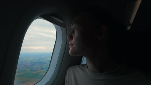 Woman looks out of airplane window during flight over landscape while clouds fill the sky