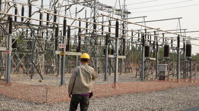 Panning right video view of electrical engineer worker holding thermal imaging camera to scan high voltage power substation switchyard equipment for maintenance inspection.