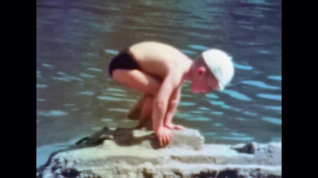 Small boy in swimsuit playing on rocky river beach on summer. Curious child climbs over stones near water, explores lake nature, enjoying outdoor adventure. Old film. Archival retro. Vintage archive