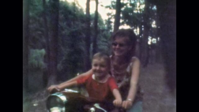 Young mother sitting on motorcycle with small son in forest. Happy, active family weekend in nature outdoor. Parent, child have fun activity together. Old film. Archival retro 1970s. Vintage archive