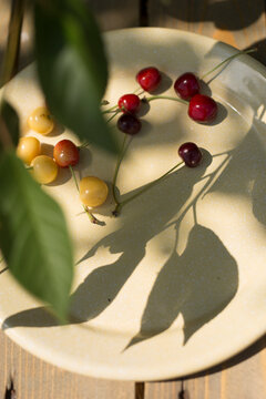 Minimalist still life of couple  red and yellow cherries with stems on a bright yellow plate in natural sunlight with hard shadows."
