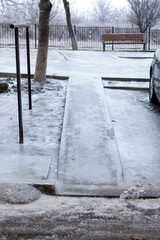 Urban sidewalk ramp covered in ice, winter trees, parked car, and empty bench in the background. Cold, overcast weather emphasizing accessibility and seasonal hazards.