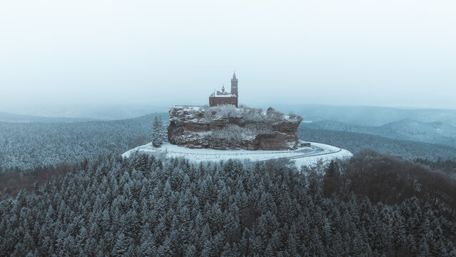 Aerial view of a snow-covered chapel atop a craggy peak, piercing the winter sky above frosted trees, Dabo, Grand Est, France.