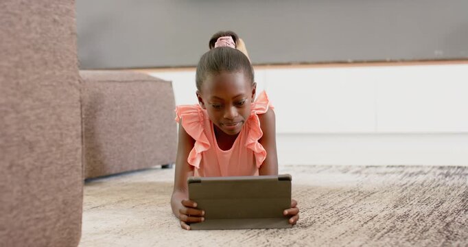 African girl in pink top lying at home, holding tablet, reacting to content, smiling then laughing
