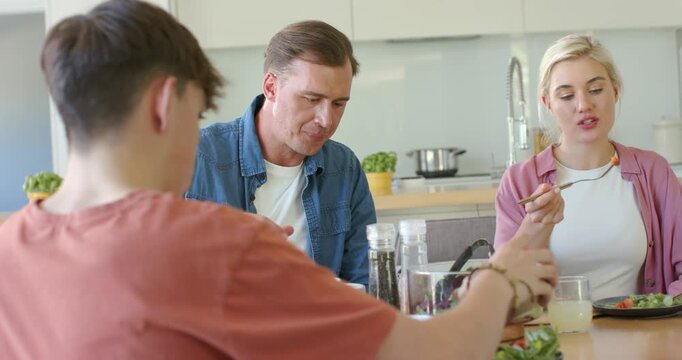 Parents and teenage son sharing salad at kitchen table, youth serving bowl, parents sipping